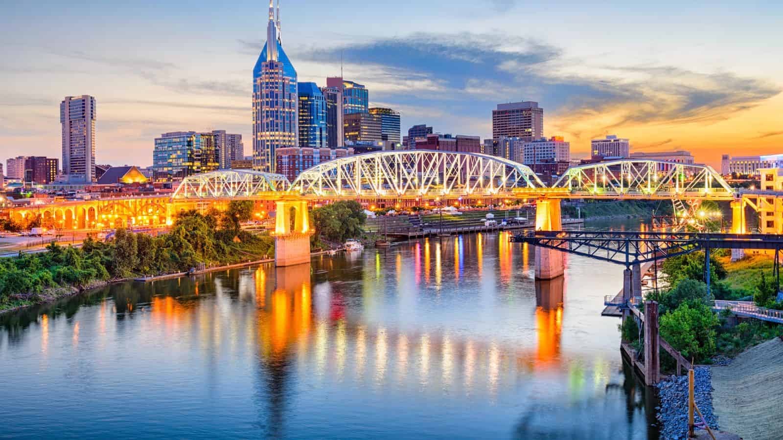 Downtown Nashville skyline at sunset with a lit bridge reflecting on the river.