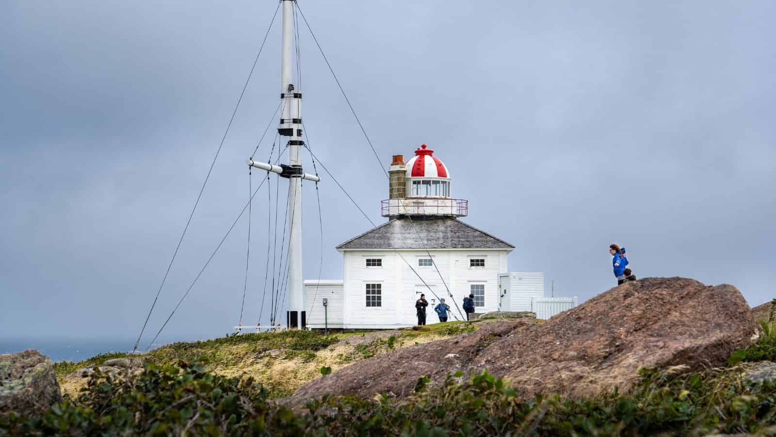 White lighthouse with red dome on a rocky hill, people walking nearby under a cloudy sky.