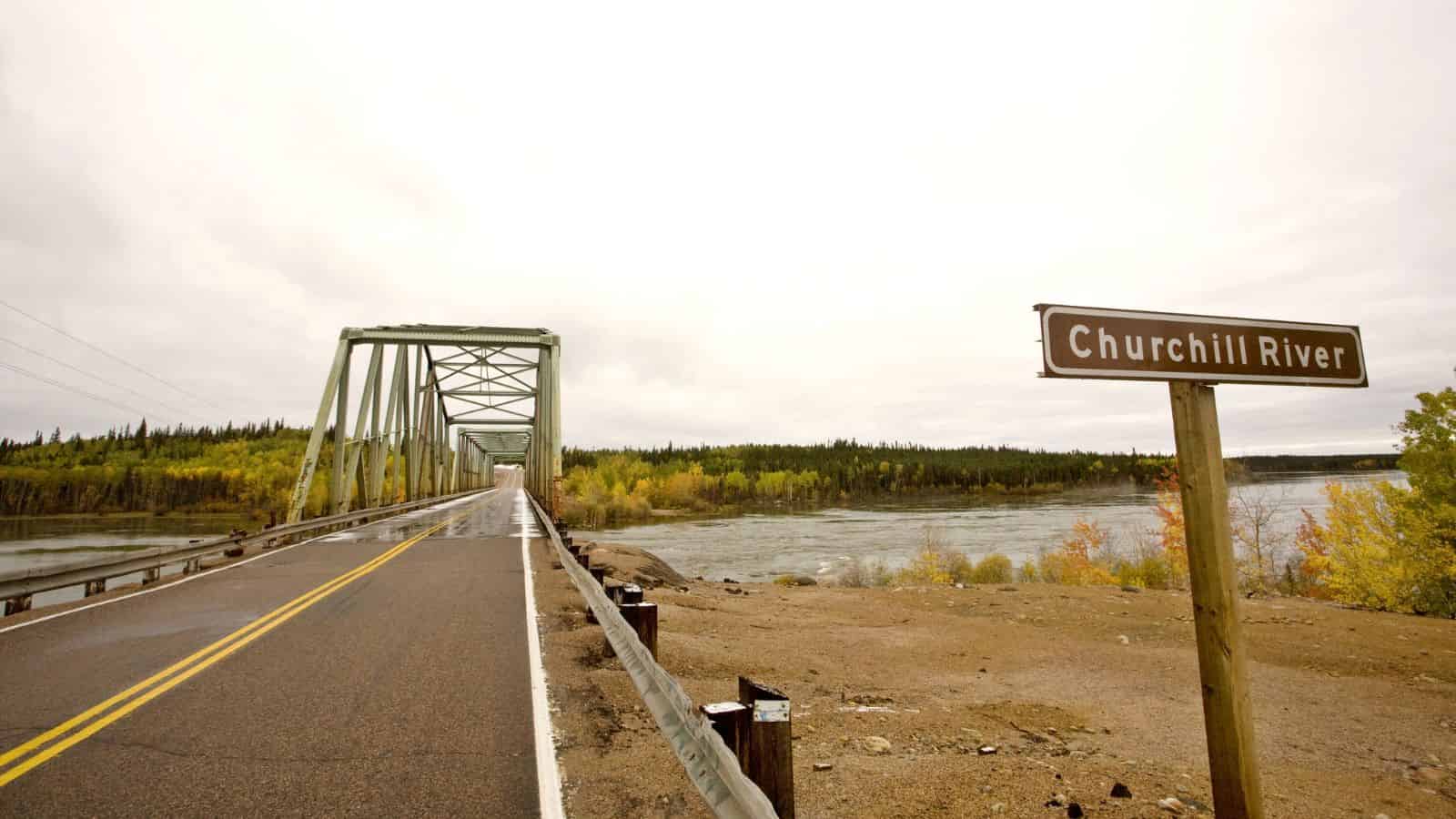 A bridge crosses the Churchill River with a sign on the right and trees in the background under a cloudy sky.