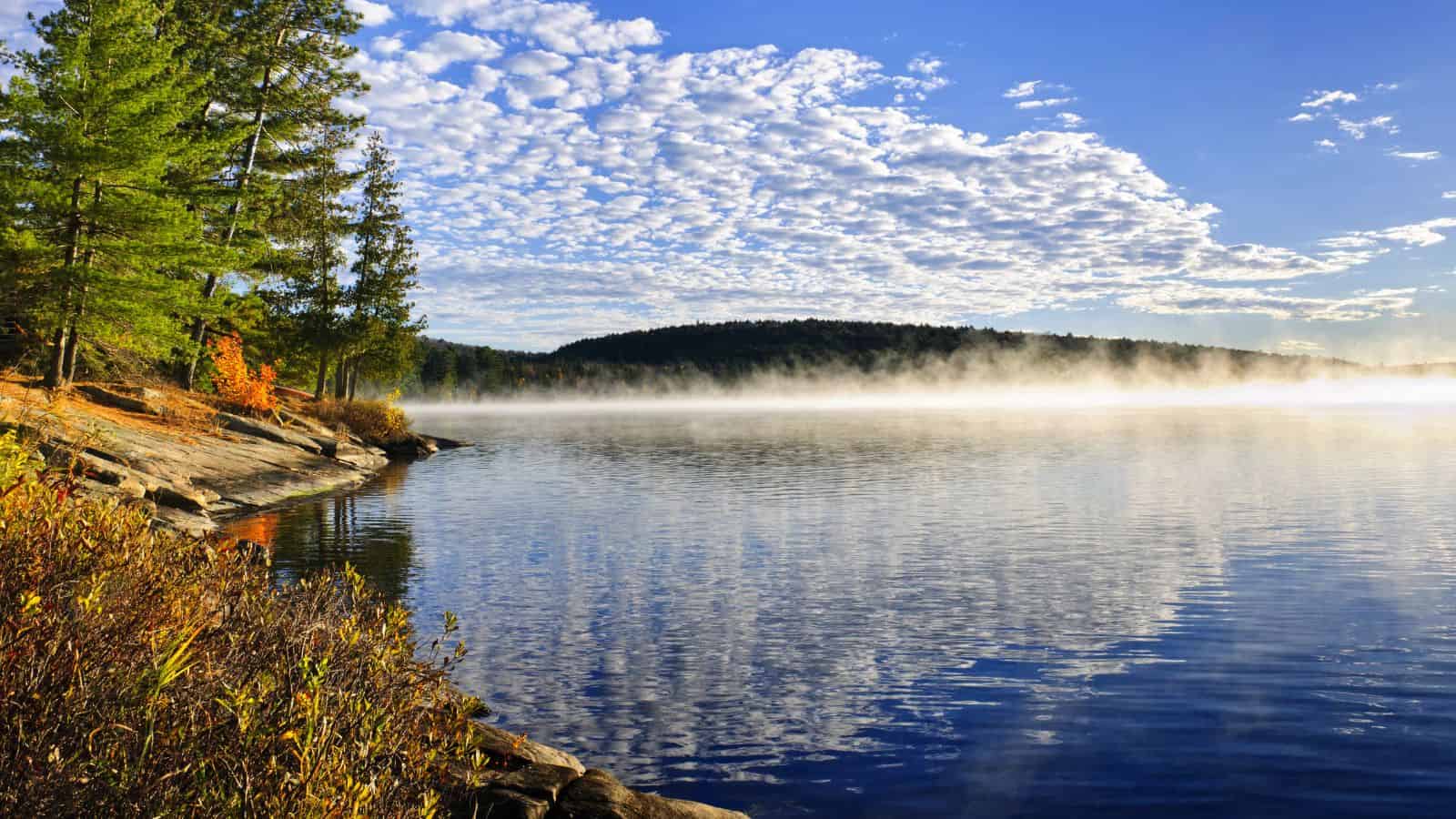 Calm lake with mist rising, surrounded by trees and rocks, under a blue sky with scattered clouds.