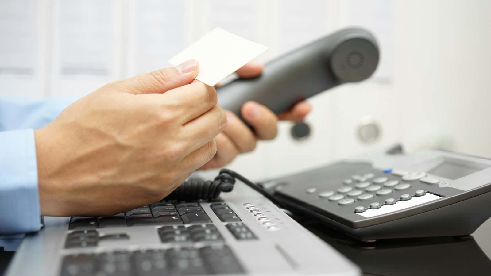 Person holding a business card and a phone handset near a keyboard and office phone on a desk, capturing old-school travel vibes and boomer memories of classic business communication.
