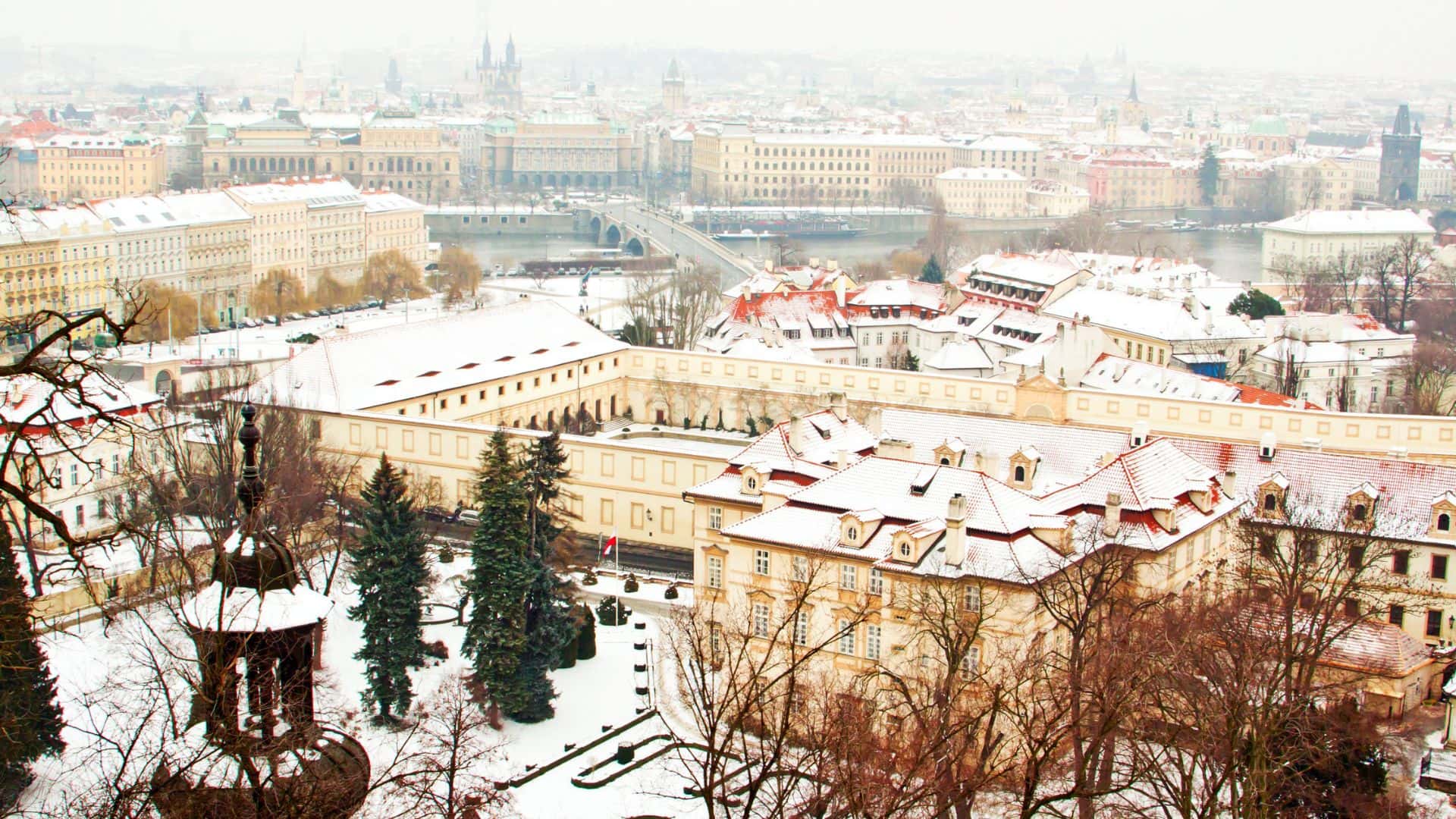 Snow-covered cityscape with historic buildings, a river, and bridges on a foggy winter day.