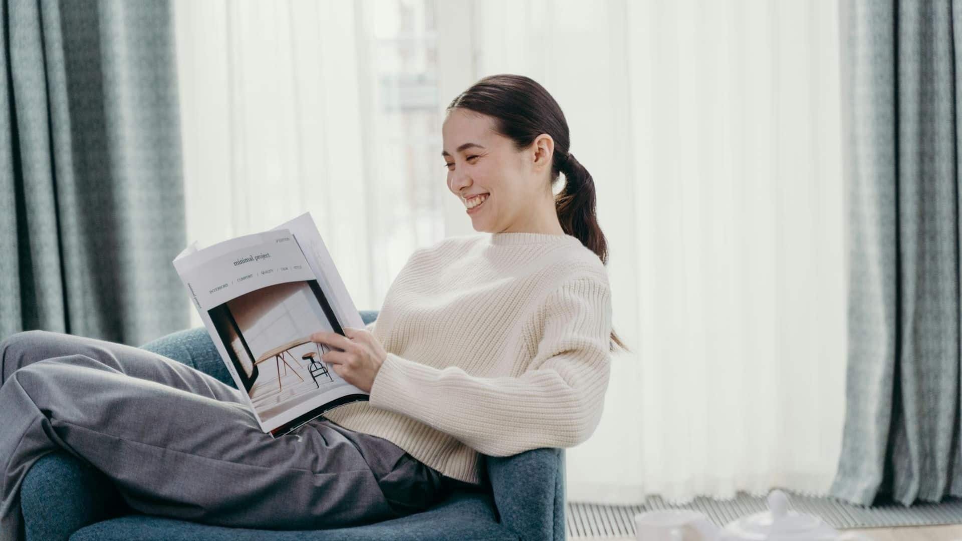 A woman sits in a chair, smiling while reading a magazine in a bright room with curtains.