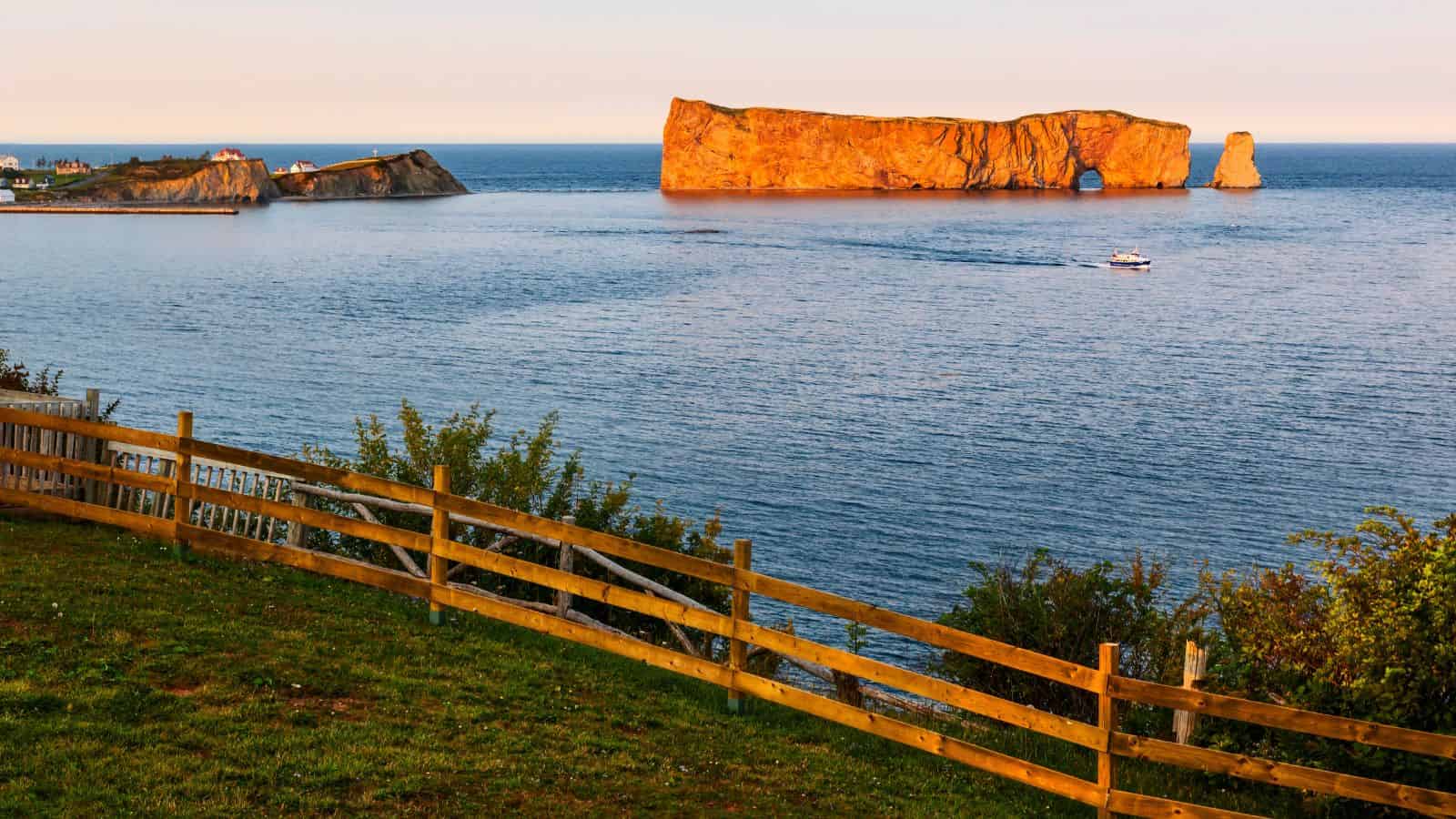 Rock formation in the sea near a coastline at sunset, with a wooden fence and greenery in the foreground.