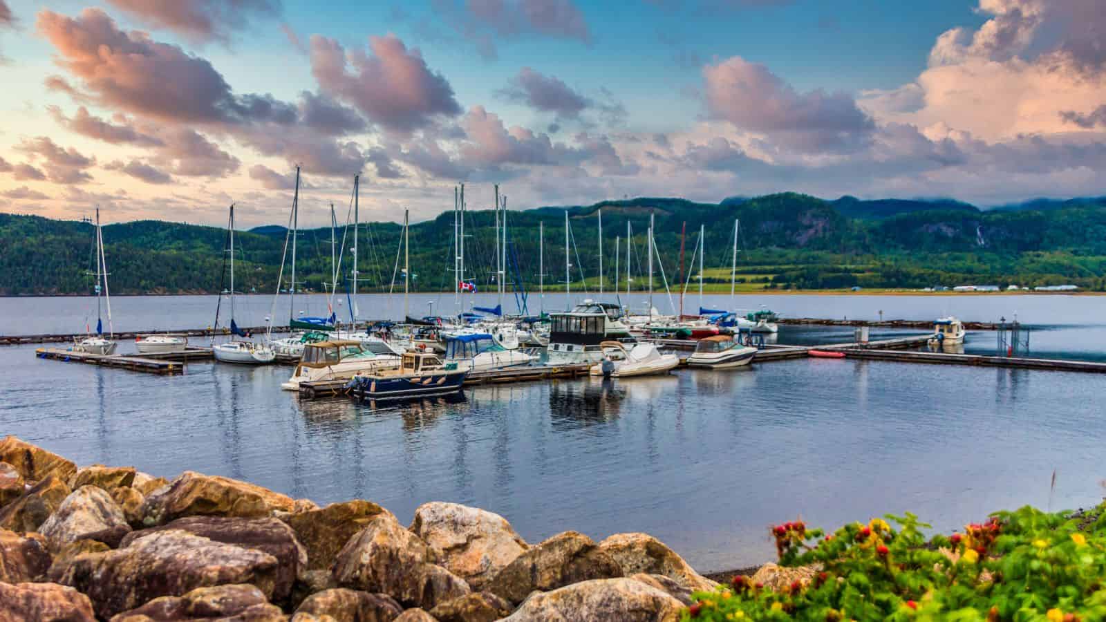 A marina with sailboats and yachts docked, surrounded by hills under a colorful sky at sunset.