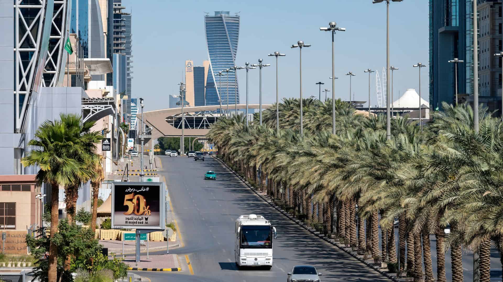 A palm-lined street in Riyadh, Saudi Arabia, with modern buildings and a distinctive leaning skyscraper.
