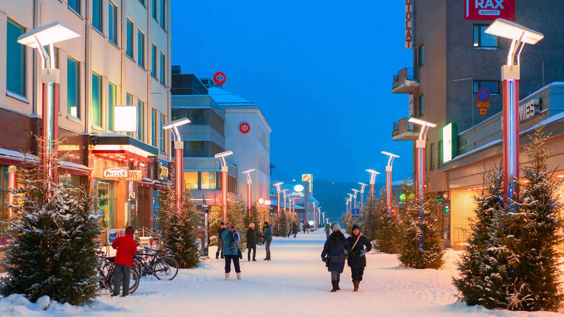 People walk down a snowy, brightly lit street lined with shops and decorated pine trees in winter.