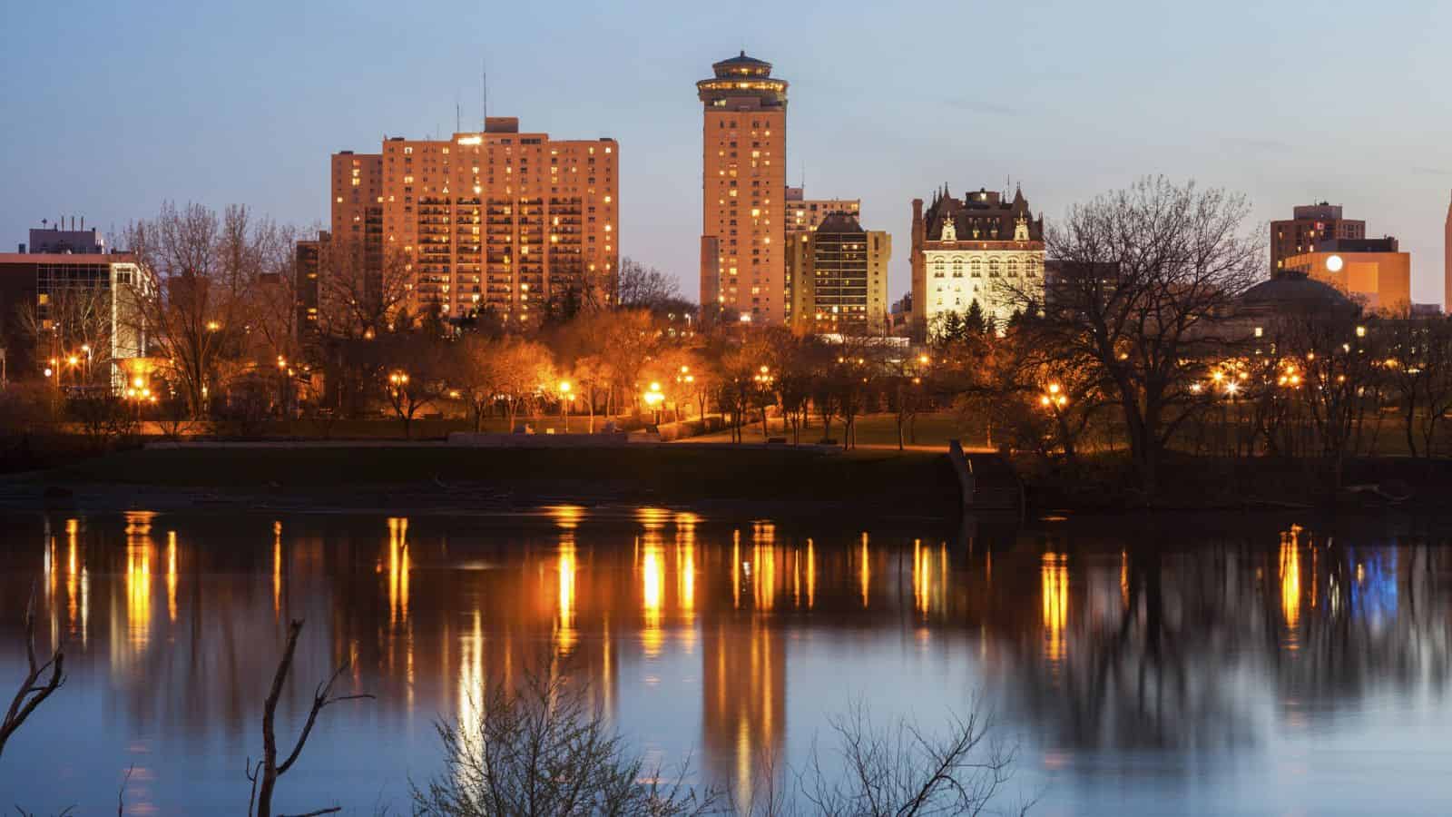City skyline at dusk with lit buildings and trees reflected in a calm river in the foreground.