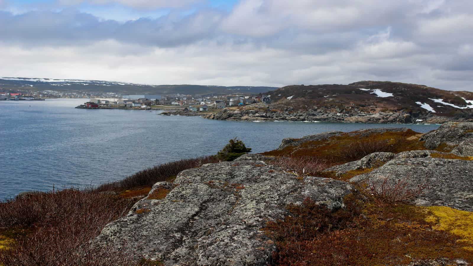 Rocky coastline with sparse vegetation, water, and a distant town under a cloudy sky.