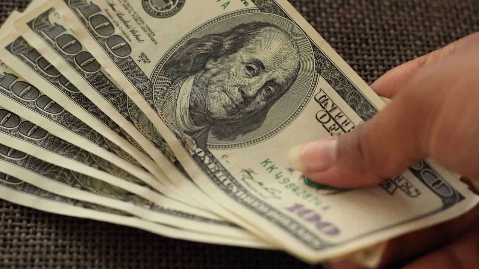 A hand holds a fan of several $100 US dollar bills on a brown textured surface.