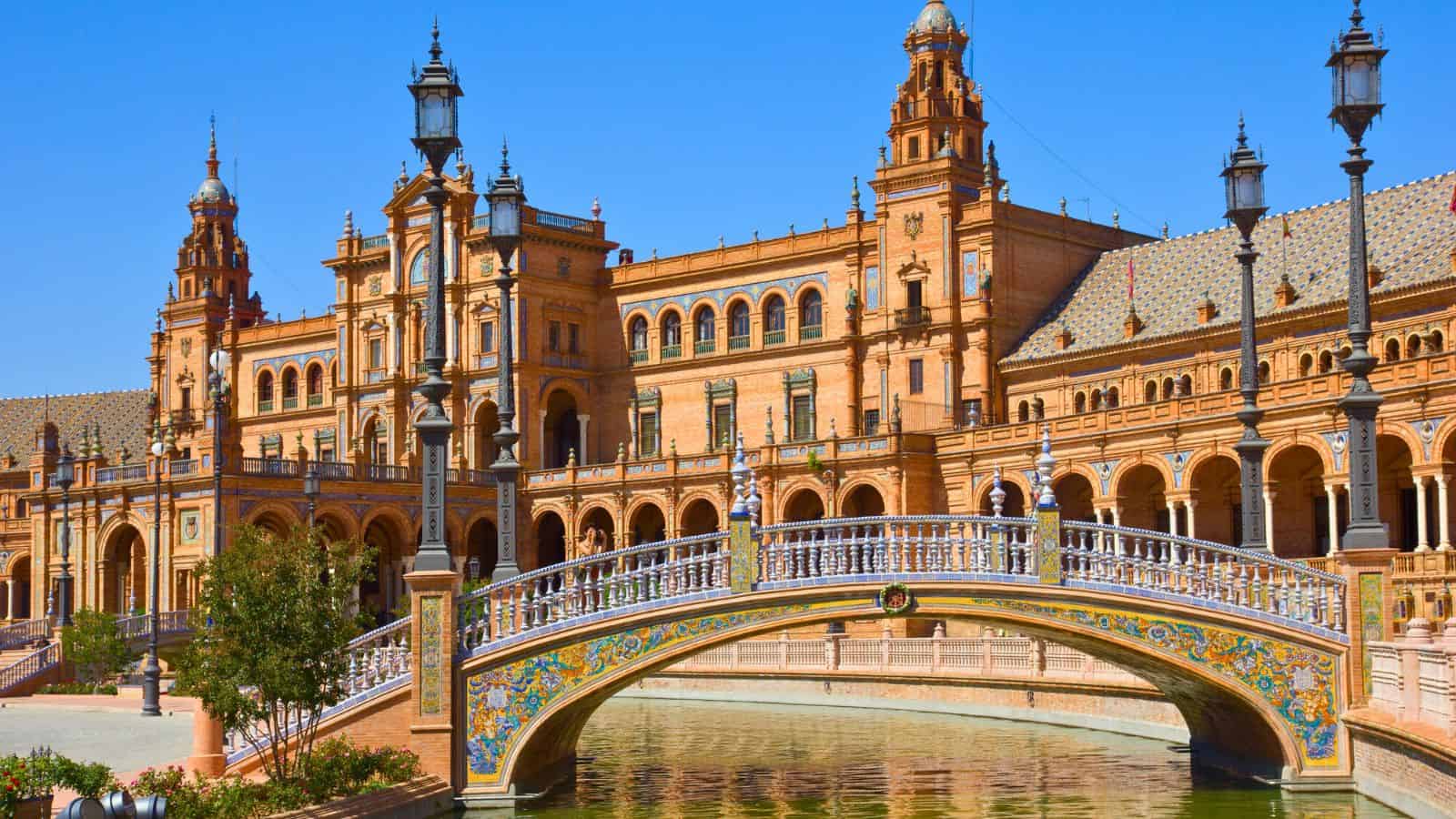 A colorful tiled bridge and ornate building at Plaza de España in Seville, Spain, under a clear blue sky.