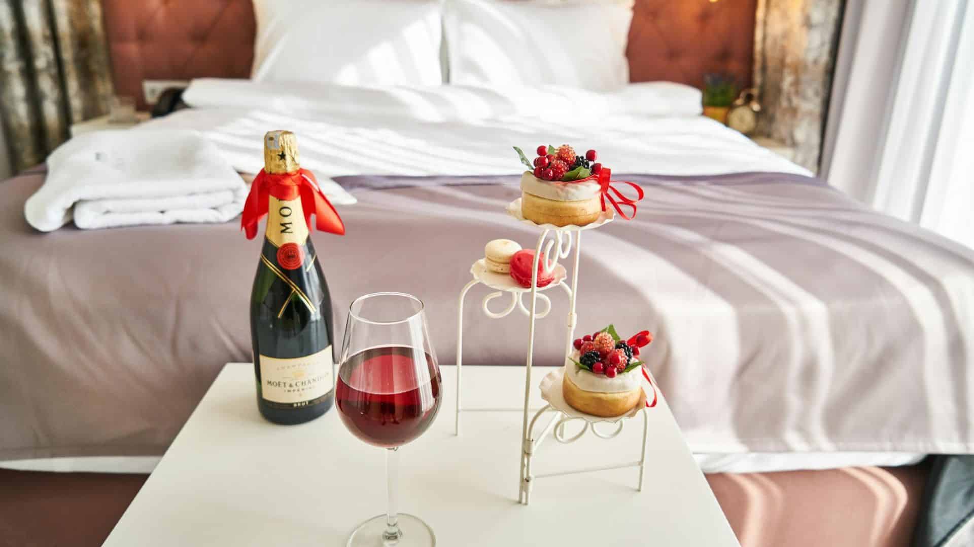 A glass of red wine, champagne, and pastries on a table in front of a cozy, neatly made bed.