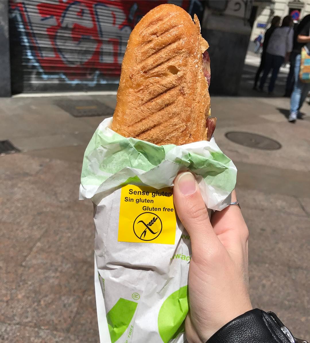 Hand holding a gluten-free sandwich wrapped in paper, set against a vibrant street with graffiti—a perfect taste of Gluten Free Barcelona.