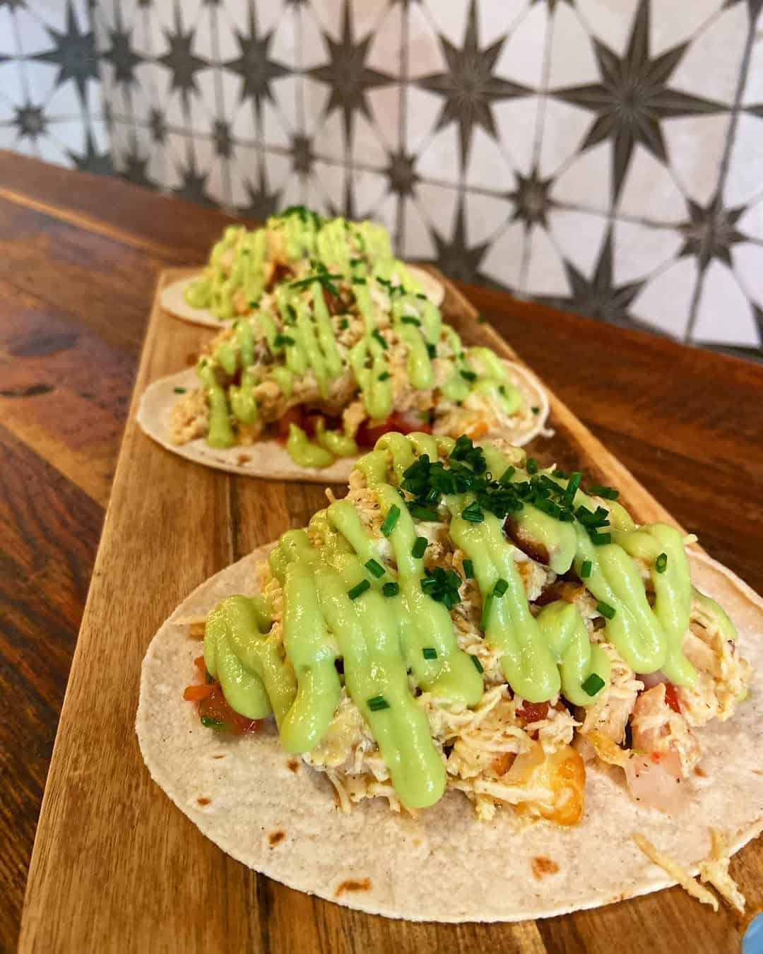 Three chicken tacos topped with green sauce and chives on a wooden board, set against a patterned tile background—a delicious Gluten Free Barcelona option.