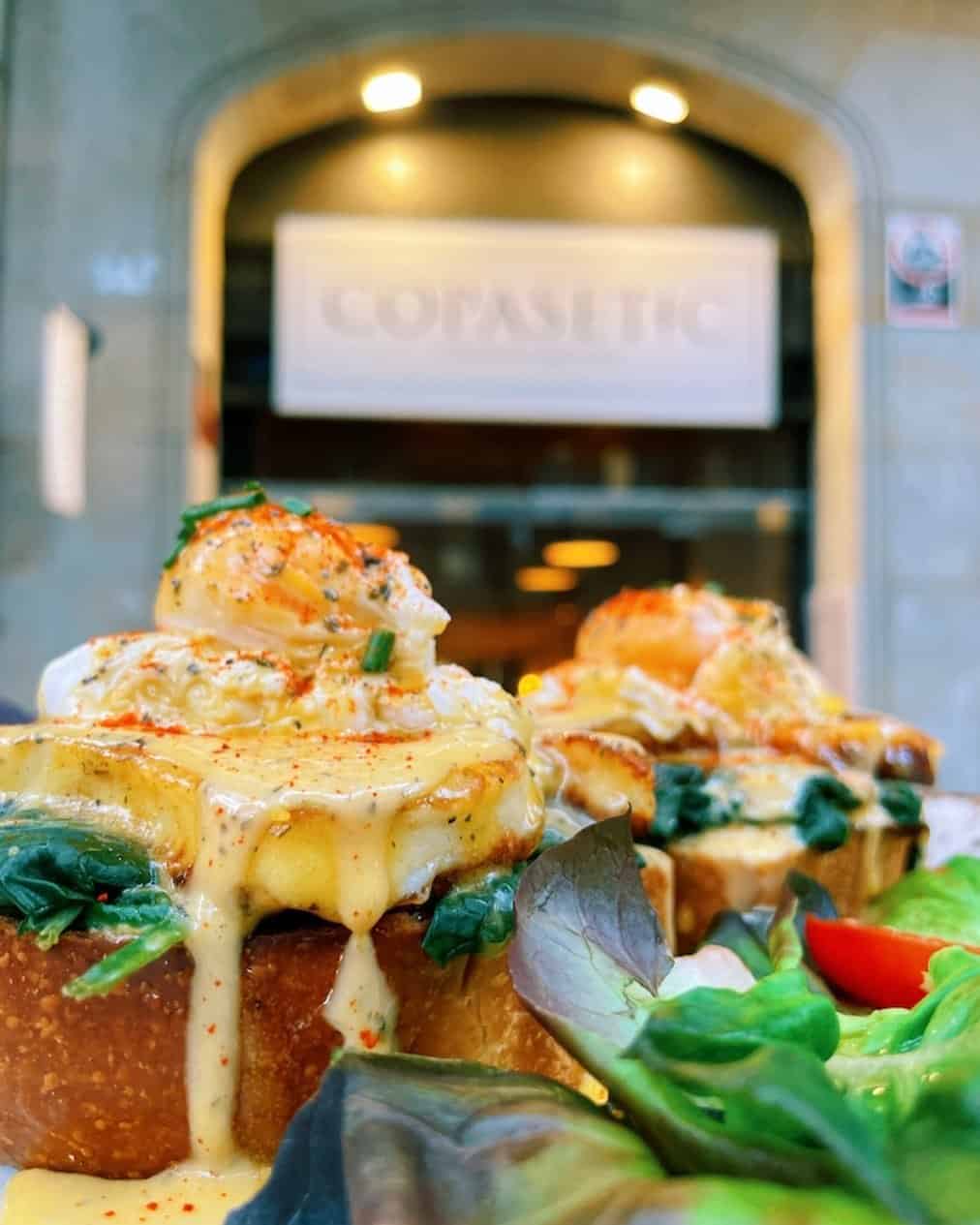 Close-up of eggs benedict with shrimp, greens, and sauce, served with salad in front of a cafe entrance—perfect for enjoying a Gluten Free Barcelona brunch.