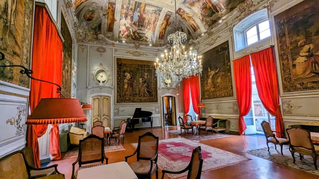 Ornate room with chandeliers, red curtains, painted ceiling, classic furniture, and wall tapestries—perfectly capturing the elegance you might find during 24 hours in Genoa, Italy.