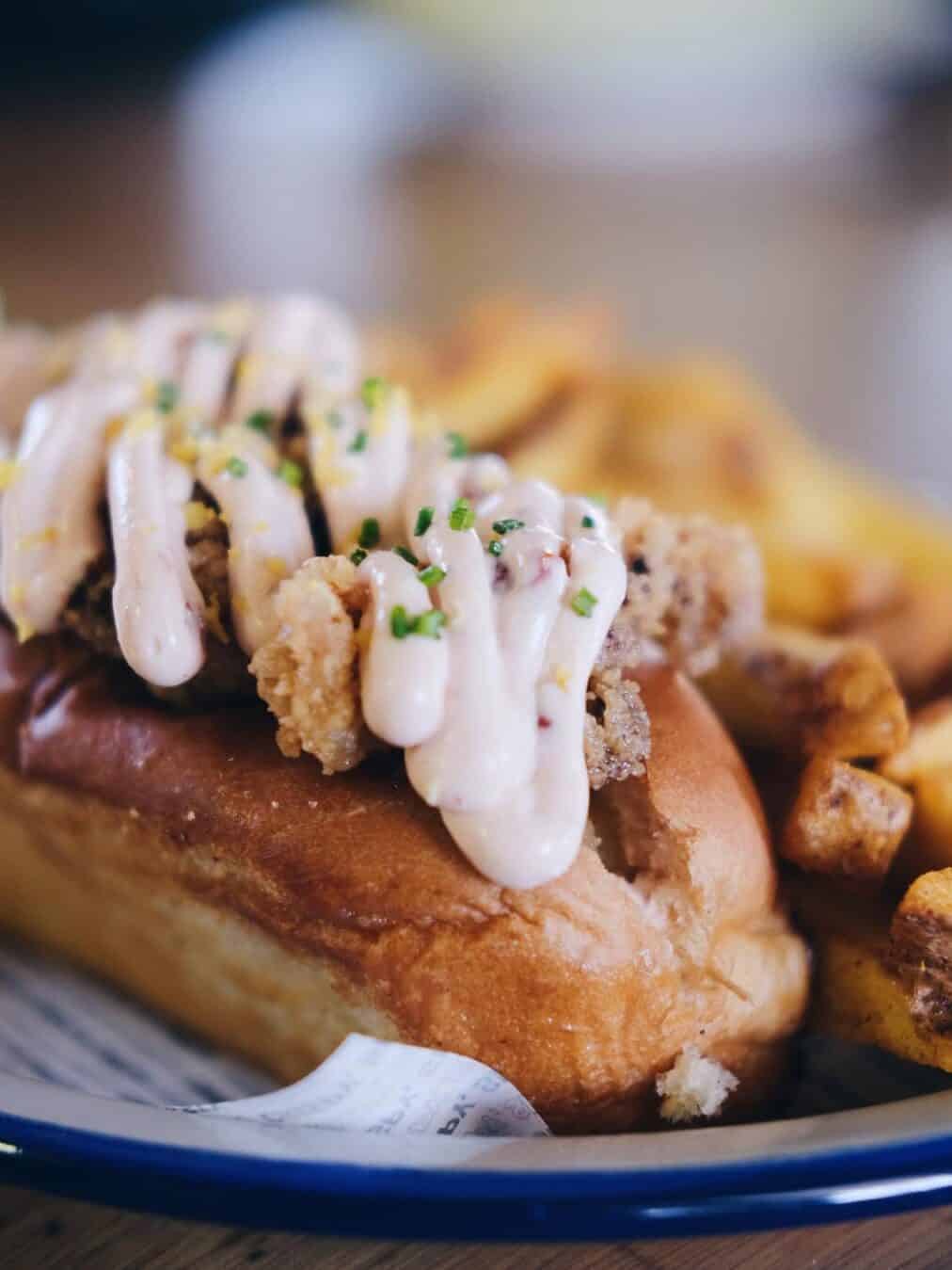 A sandwich roll with fried chicken, creamy sauce, chives, and a side of seasoned fries on a plate—perfect for those seeking Gluten Free Barcelona flavors.