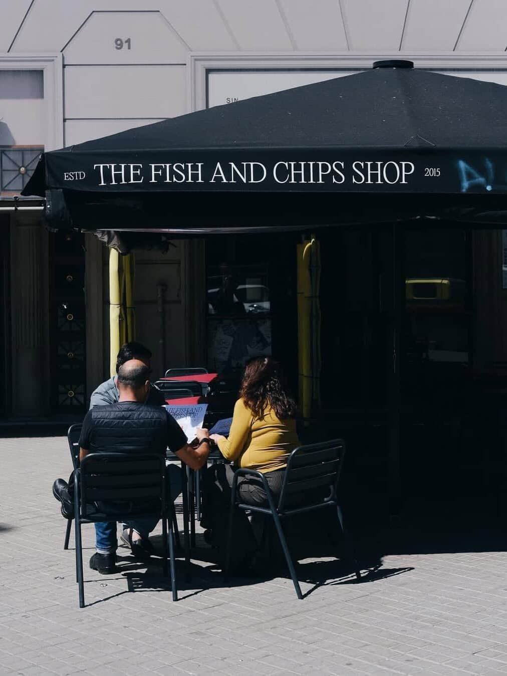 Three people sit at a table under a black umbrella labeled "The Fish and Chips Shop" on a sunny day, enjoying their meal at this popular spot known for its tasty gluten free Barcelona options.