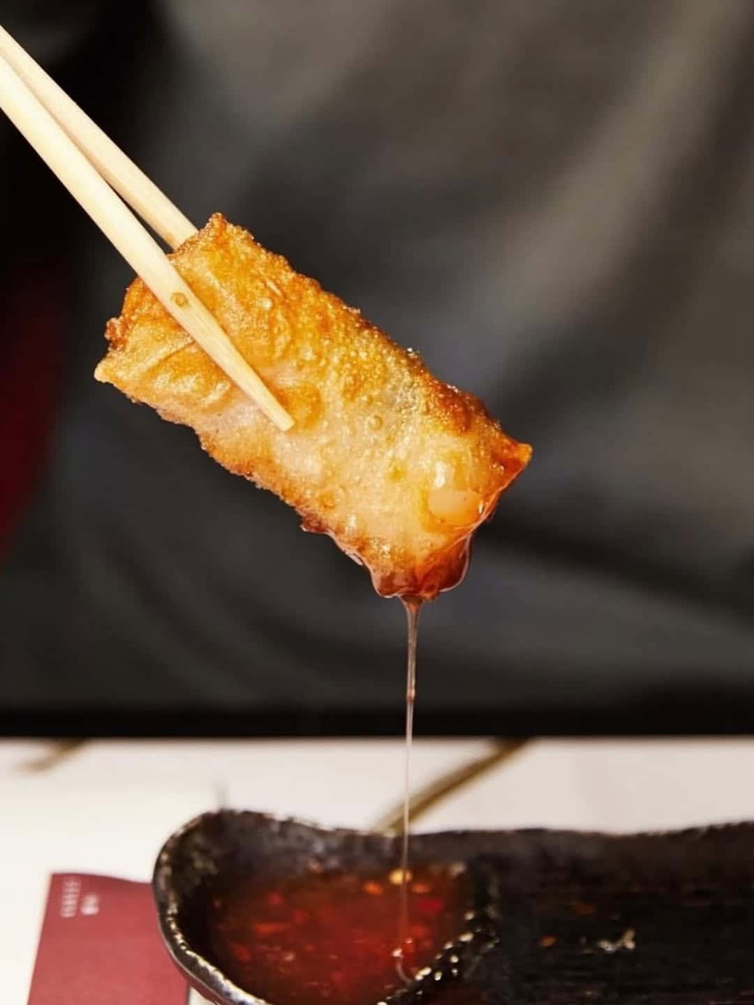 A fried spring roll held by chopsticks, dipping into a bowl of red sauce with sauce dripping—perfect for those seeking Gluten Free Barcelona dining options.
