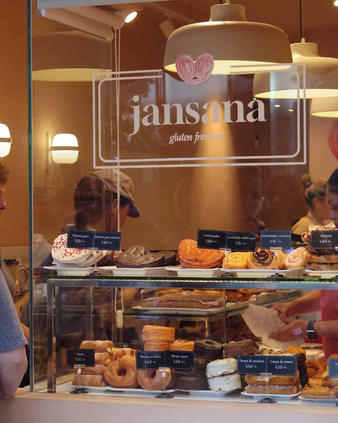 Bakery display case with assorted pastries and gluten-free signs, viewed through a window marked “jansana gluten freedom”—a delicious glimpse of Gluten Free Barcelona delights.