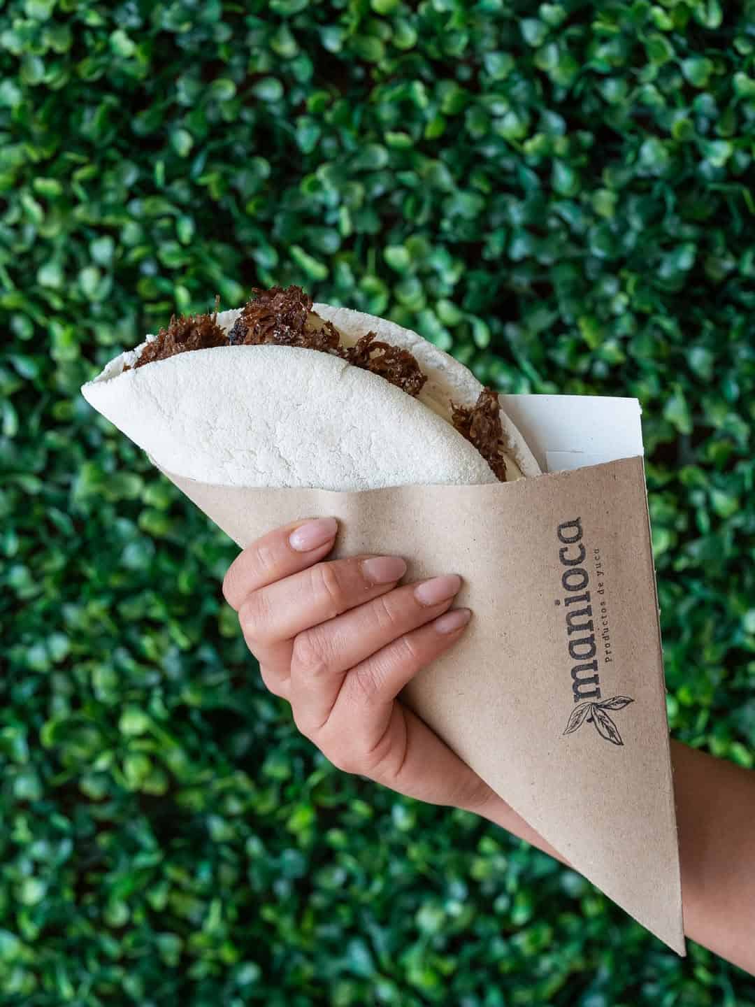 A hand holds a folded tapioca wrap filled with shredded meat, against a green leafy background—perfect for anyone seeking gluten free Barcelona eats.