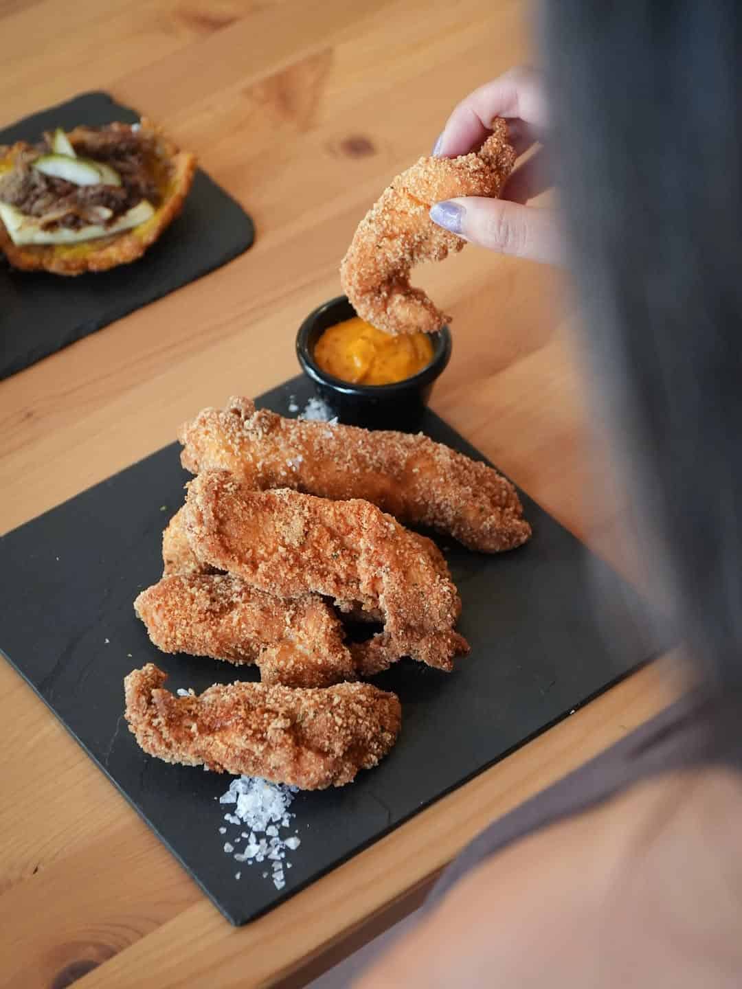 A person dips a crispy, gluten free chicken tender into orange sauce, with more tenders and sea salt on a slate plate—perfect for Gluten Free Barcelona food lovers.