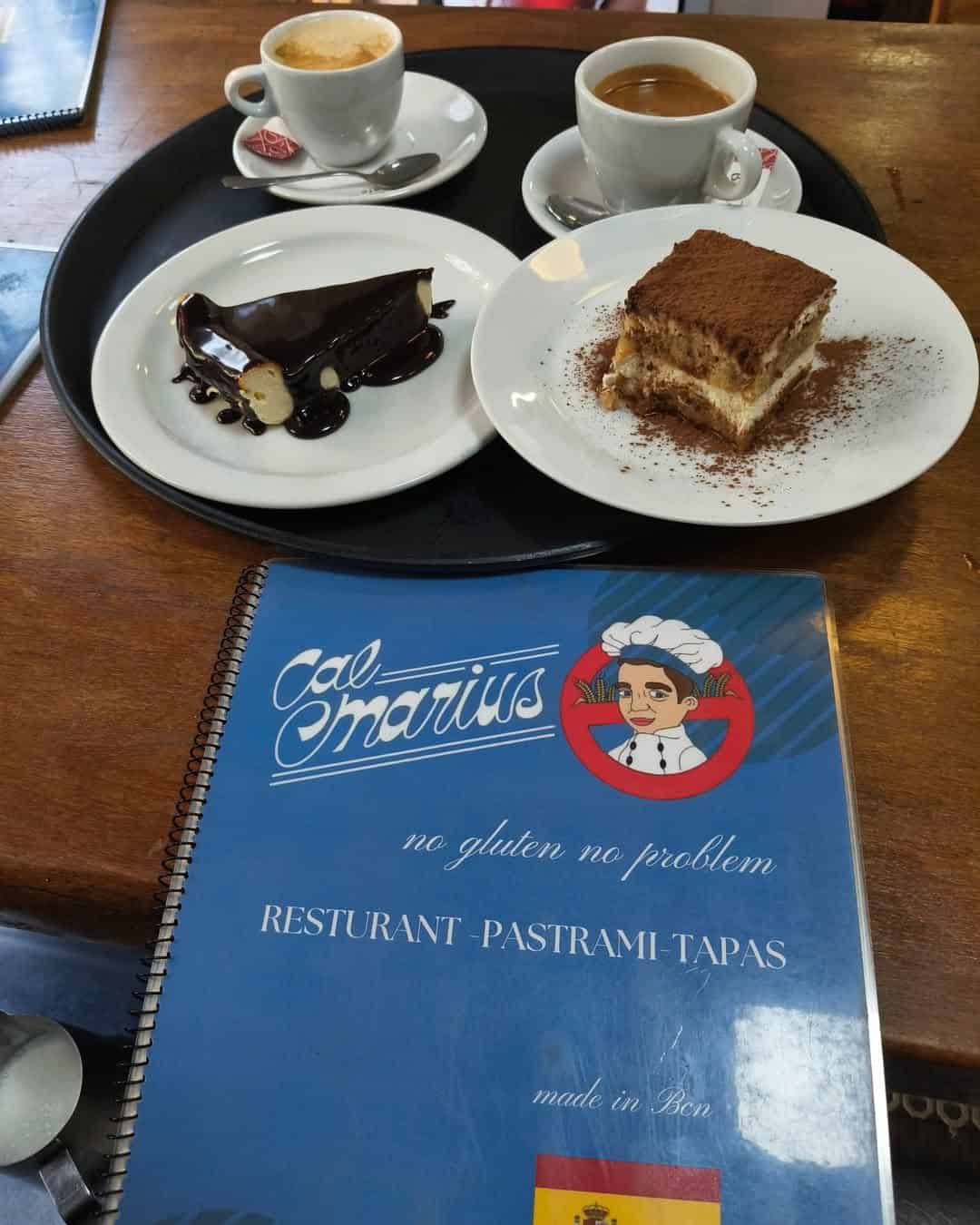 Two coffees, a slice of tiramisu, a chocolate dessert, and a blue restaurant menu on a table—perfect for discovering Gluten Free Barcelona treats.