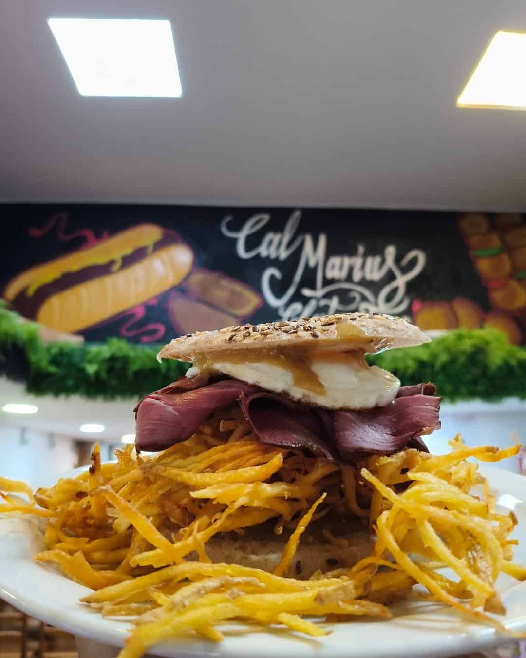 A sandwich with pastrami, cheese, sauce, and crispy fries on a plate, with a deli sign in the background—perfect for anyone seeking Gluten Free Barcelona options.