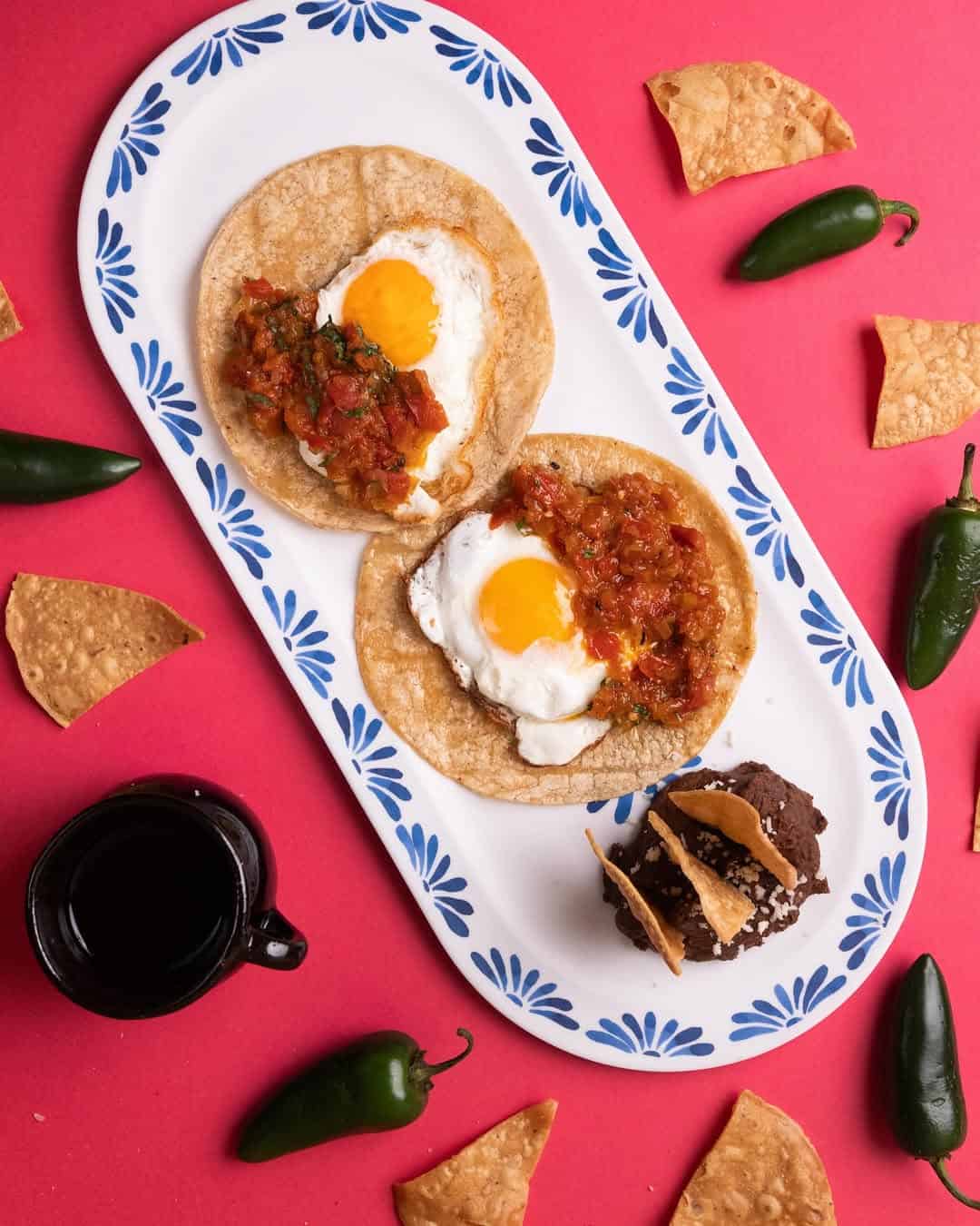 Two huevos rancheros on tortillas with salsa, a dessert, coffee, chips, and jalapeños on a red background—perfect for anyone seeking Gluten Free Barcelona options.