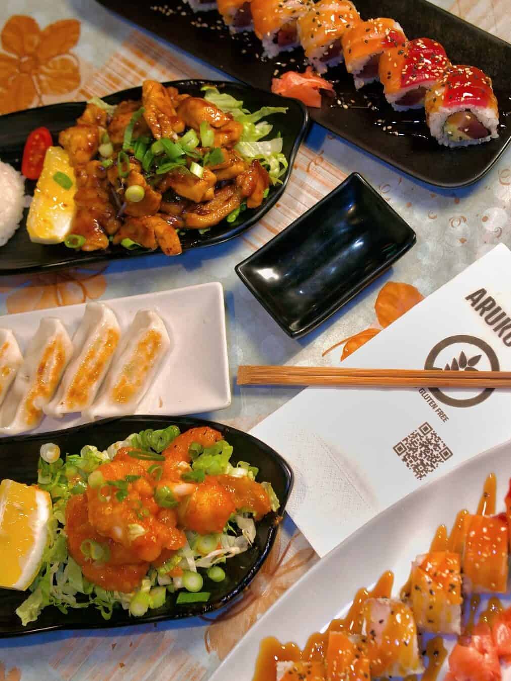 A table set with sushi rolls, dumplings, and Asian dishes, along with chopsticks and a restaurant menu, offers a glimpse of Gluten Free Barcelona dining options.