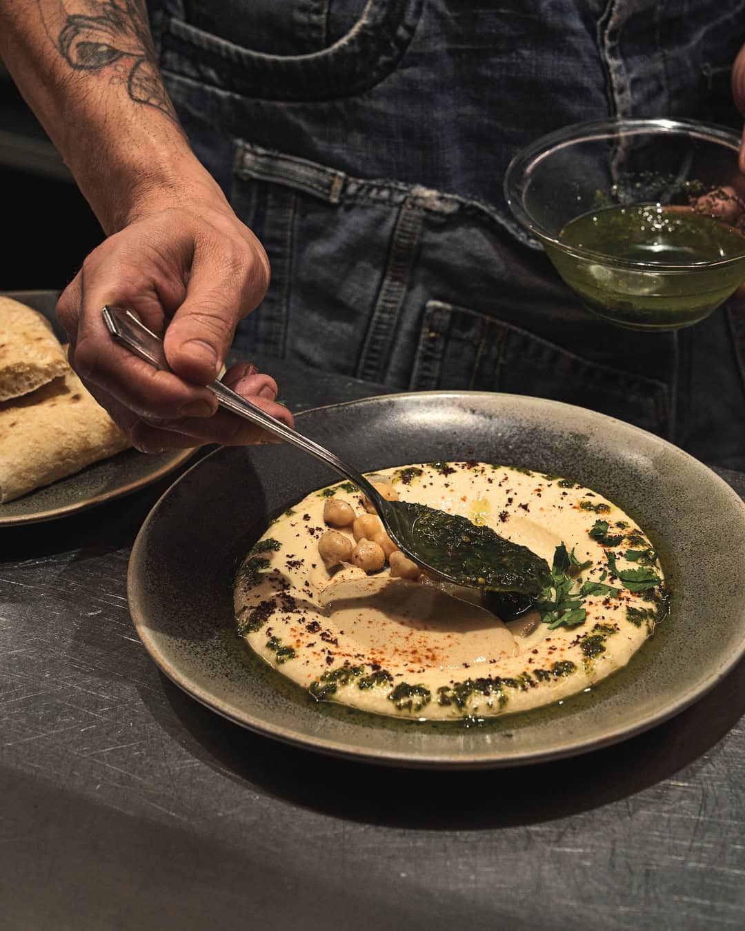 A person spoons green sauce onto a plate of hummus garnished with chickpeas and herbs, perfect for those seeking Gluten Free Barcelona dining options.