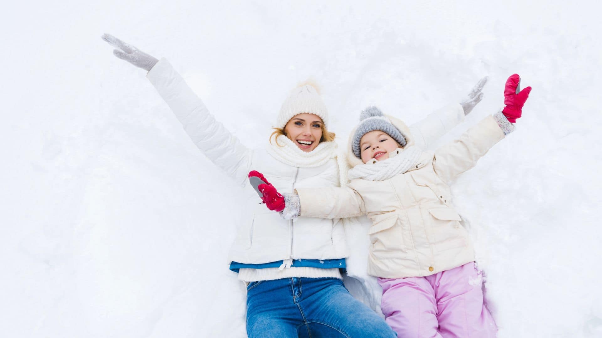 Woman and child lying in snow with arms outstretched, smiling and dressed in winter clothes.