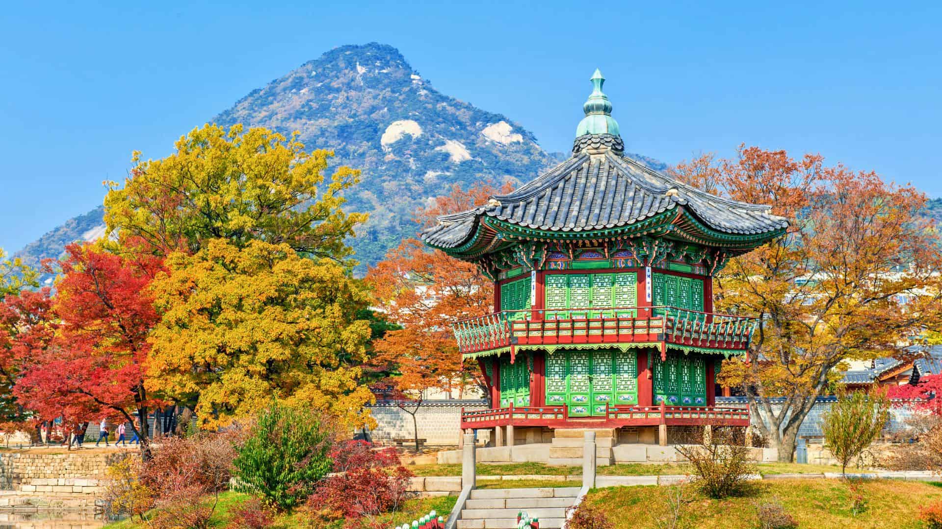 Traditional Korean pavilion with colorful autumn trees and a mountain in the background under a clear blue sky.