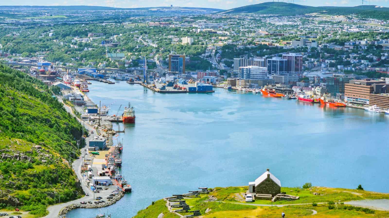 A view of a harbor with ships, buildings, and green hills under a partly cloudy sky.