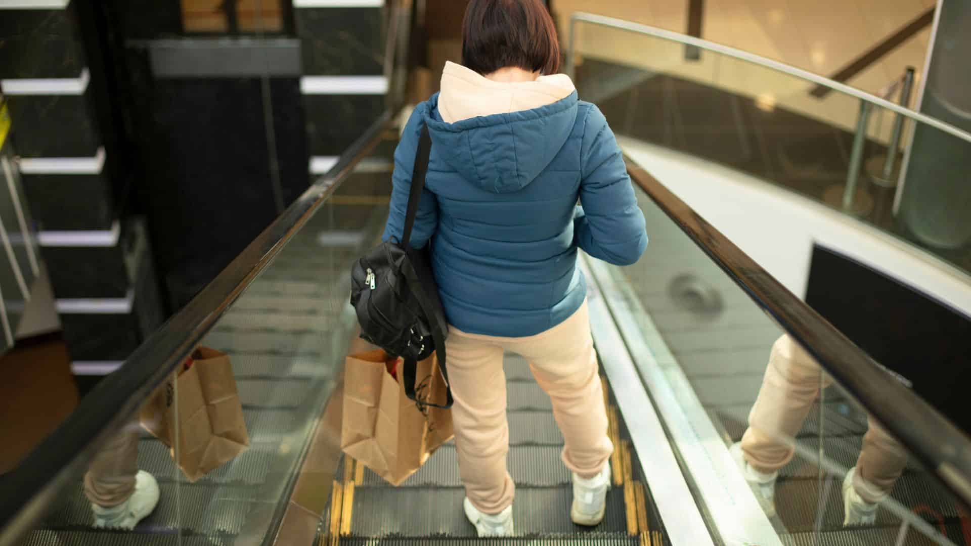 Person with a backpack and shopping bags riding down an escalator inside a mall or shopping center.
