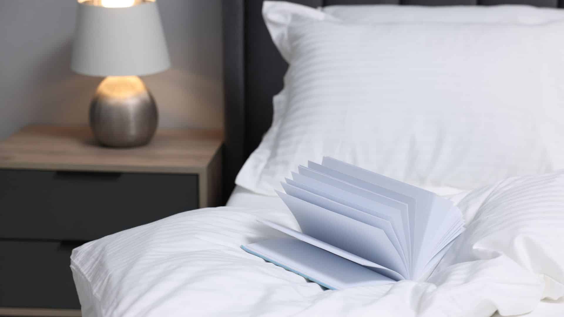 A book lies open on a neatly made bed next to a bedside table with a lamp.