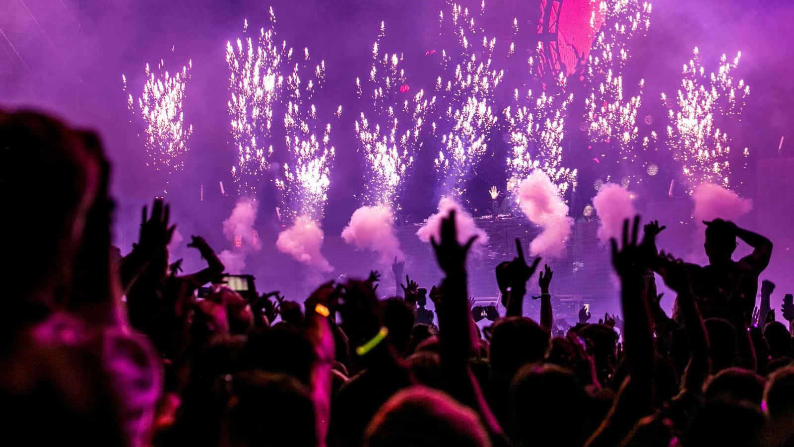 Crowd cheering at a concert with purple lights, fireworks, and smoke effects on stage.