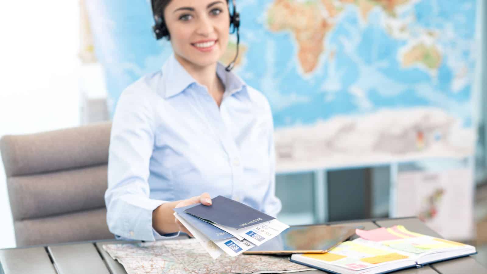 Smiling travel agent with headset hands over passports and tickets, with a map and documents on the desk.