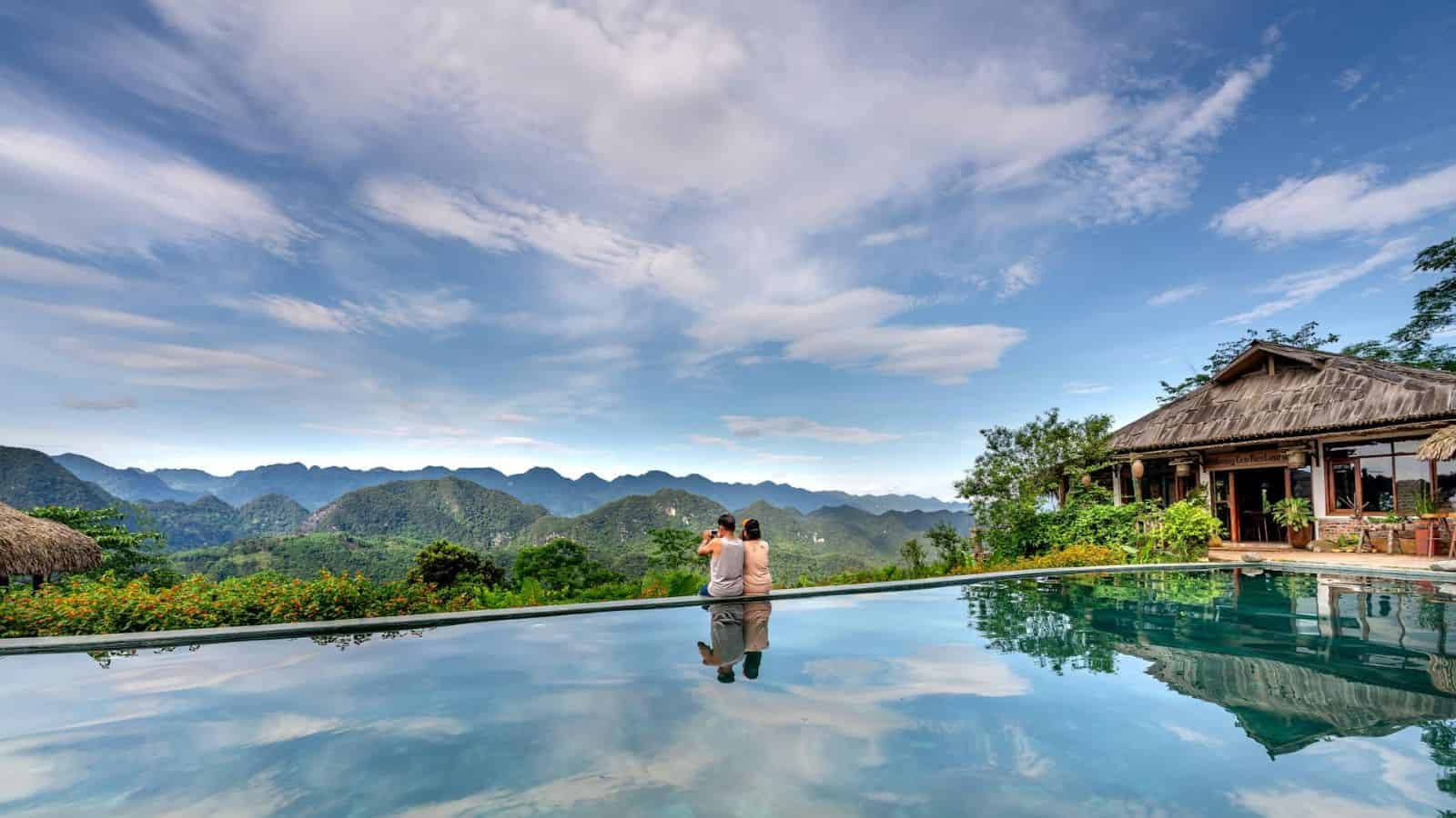 Two people sit by an infinity pool with mountain views, a rustic building, and a bright blue sky with clouds.