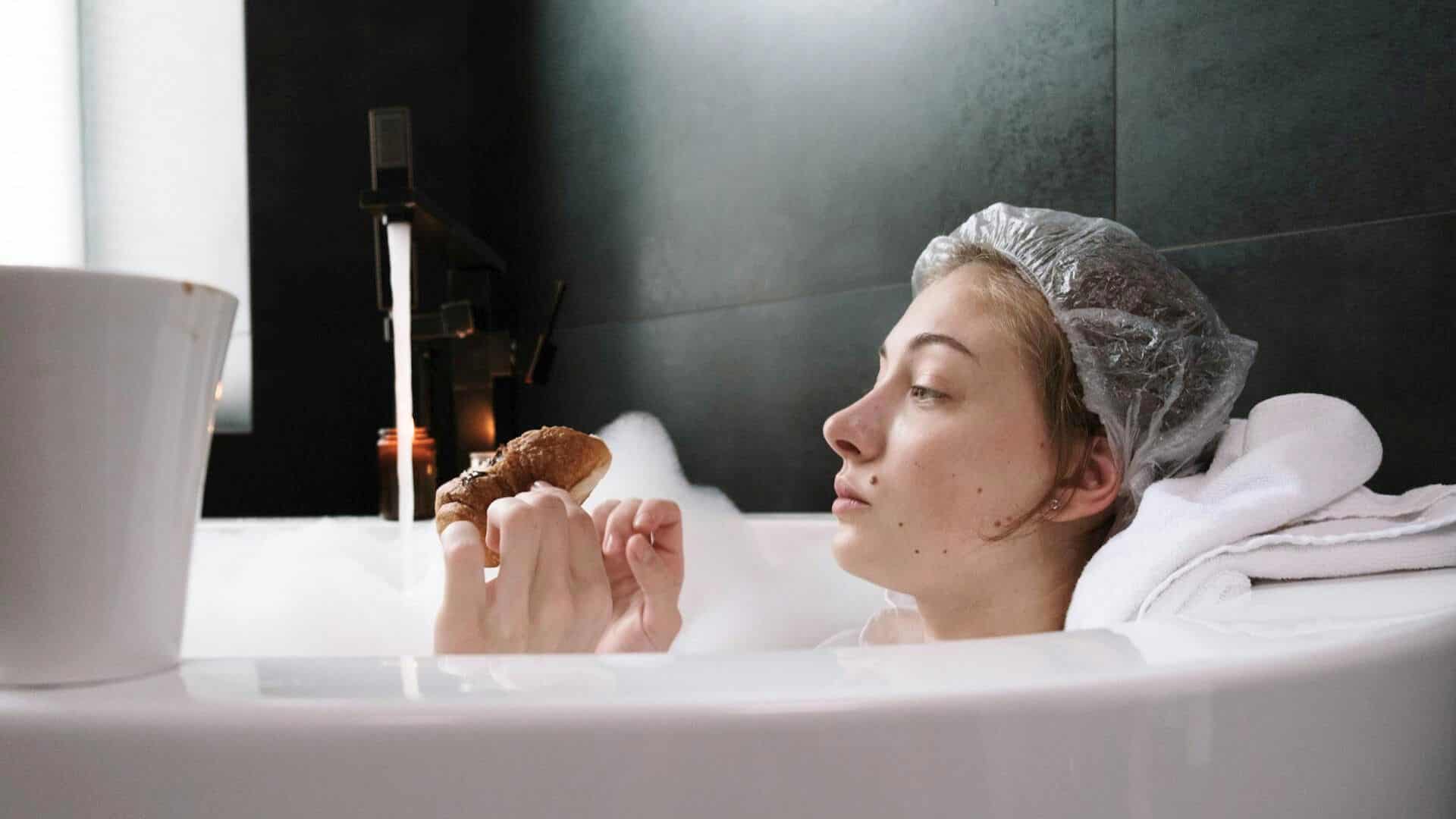 A person in a bubble bath, wearing a shower cap, holds a pastry and looks at it thoughtfully.