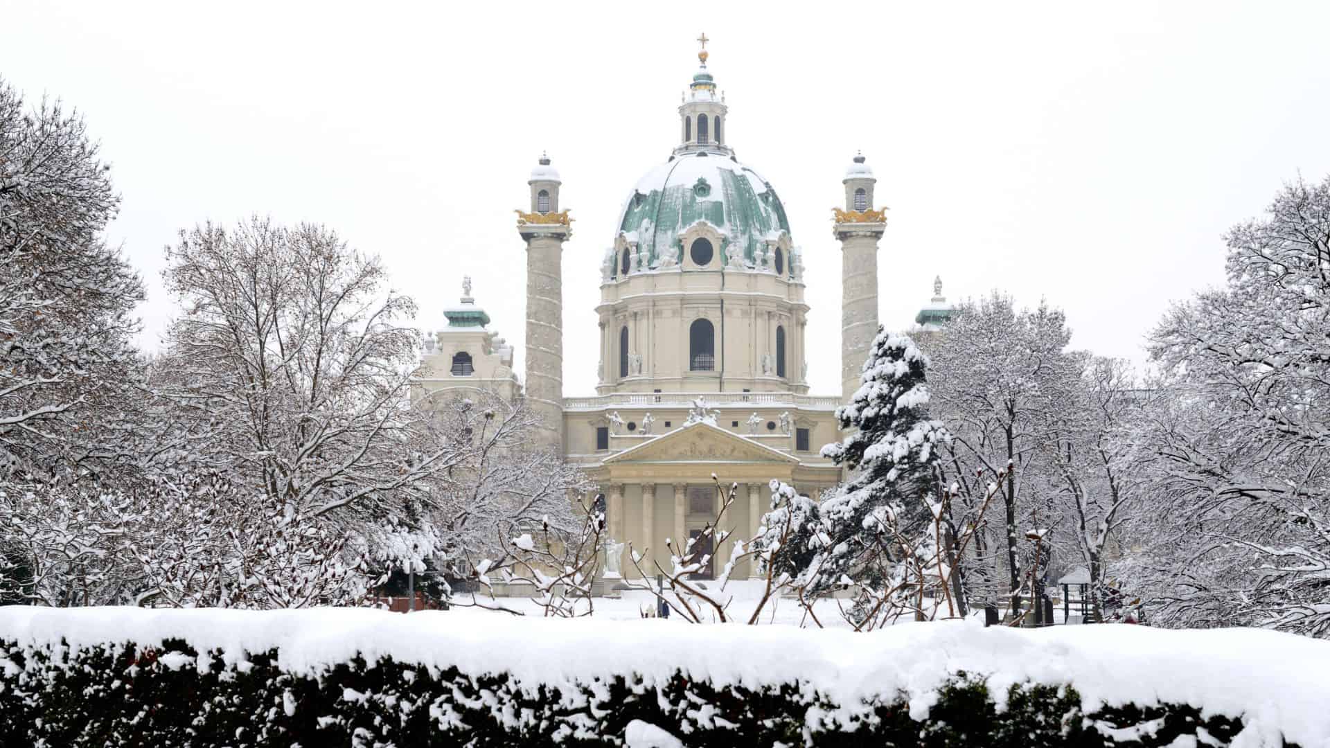 Snow-covered trees and bushes in front of a grand domed cathedral building in winter.