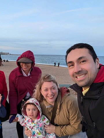 Five adults and two children smile together on a beach in Barcelona, with a city building and ocean in the background—capturing the joy of 4 days in Barcelona.