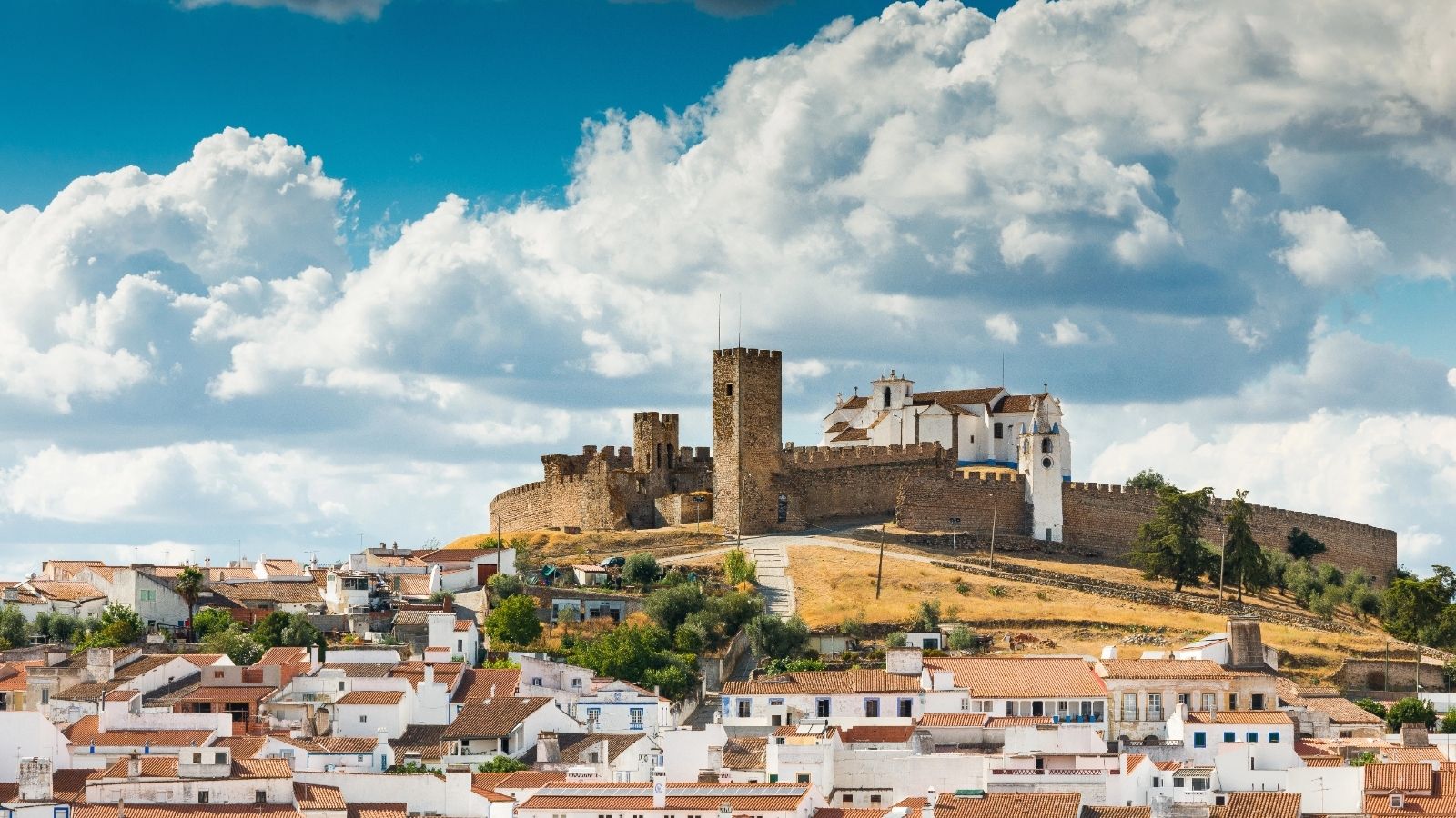 A medieval castle on a hill overlooks white houses with red roofs under a blue sky with clouds.