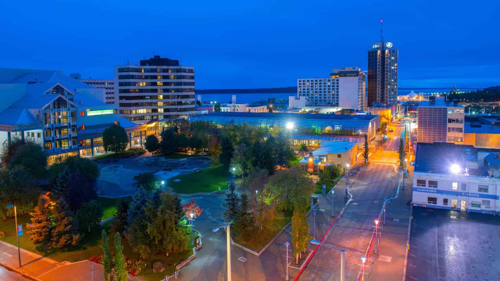 A cityscape at dusk with lit buildings and trees lining a park and a street.