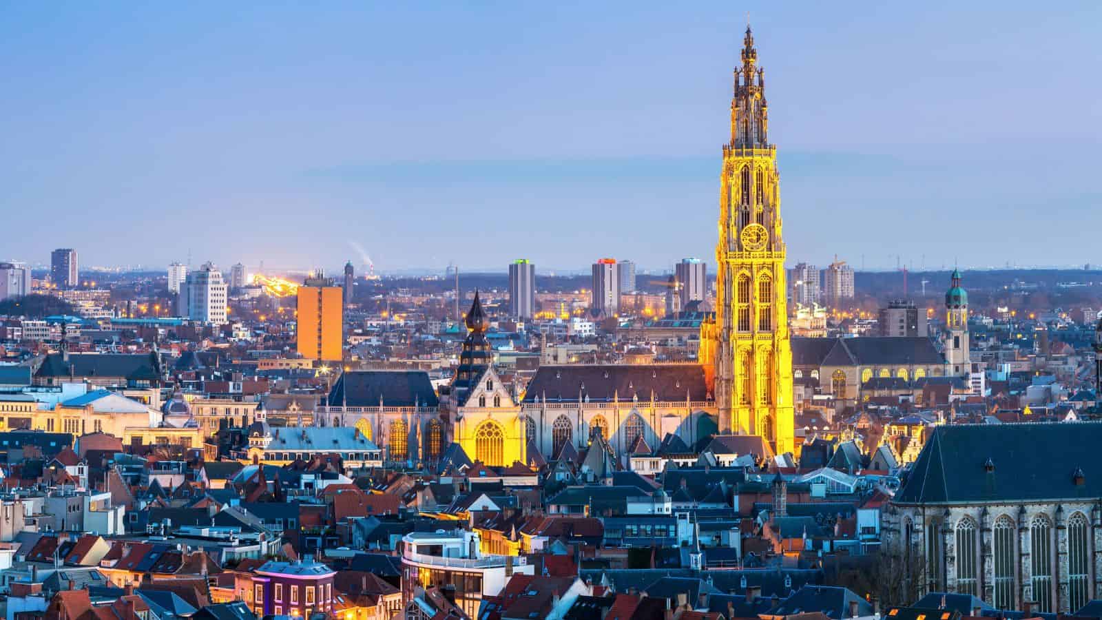 A cityscape at dusk with a tall, illuminated Gothic cathedral tower rising above rooftops and modern buildings.