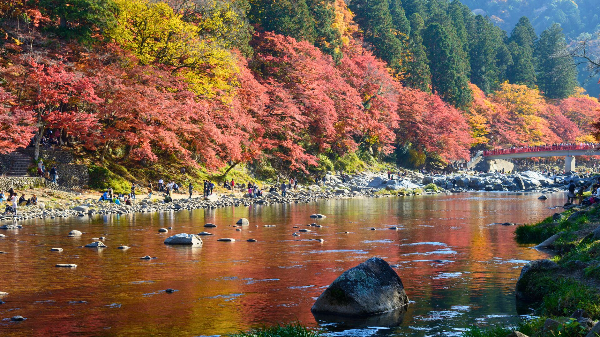 A river reflects vibrant autumn trees with red and orange leaves; people walk along the rocky shore.