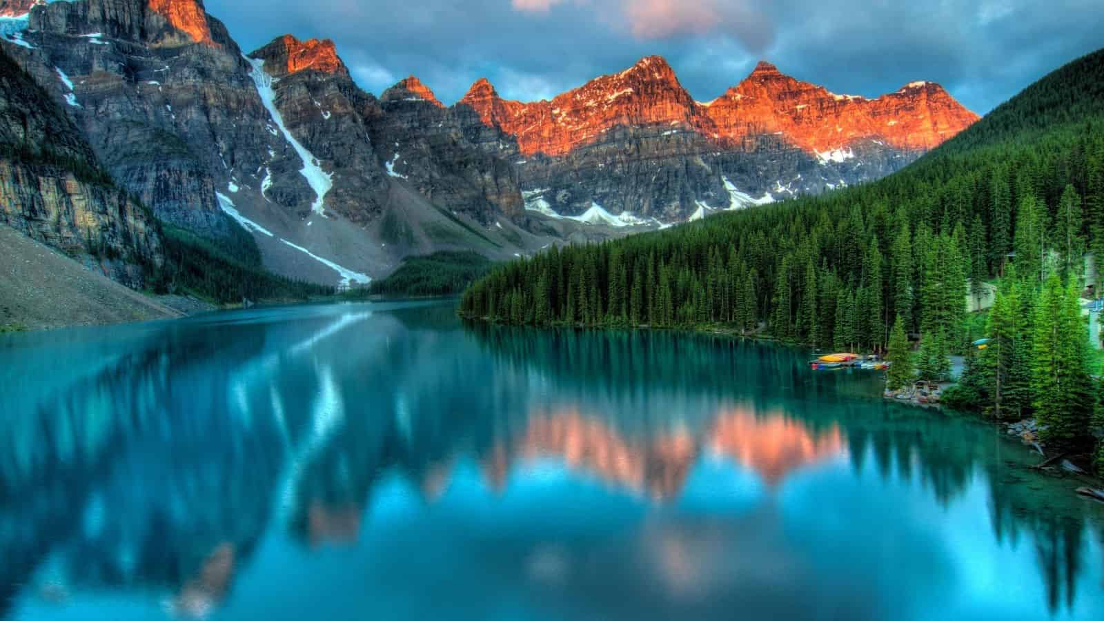 Snow-capped mountains at sunrise reflected in a calm blue lake surrounded by dense pine forest.