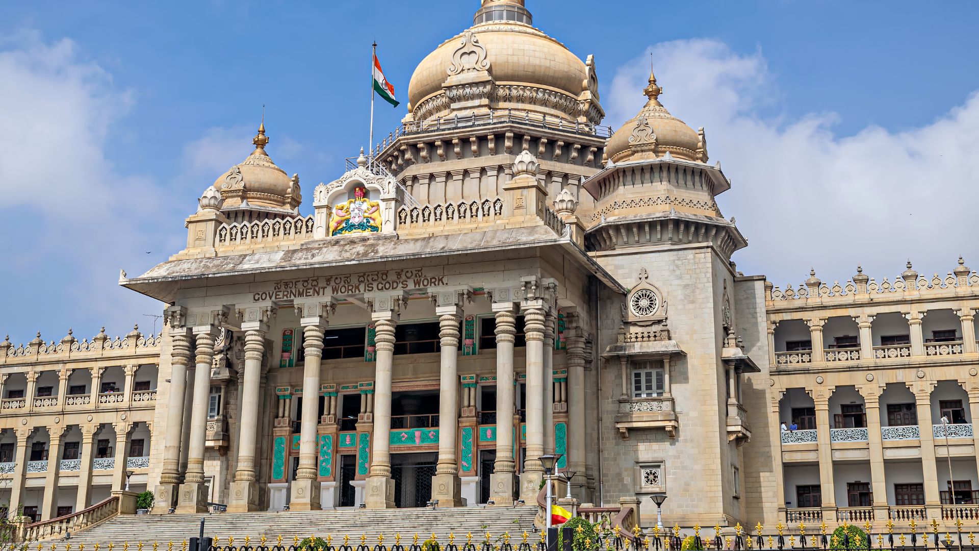 A grand government building with domes, columns, and flying Indian flags under a blue sky.