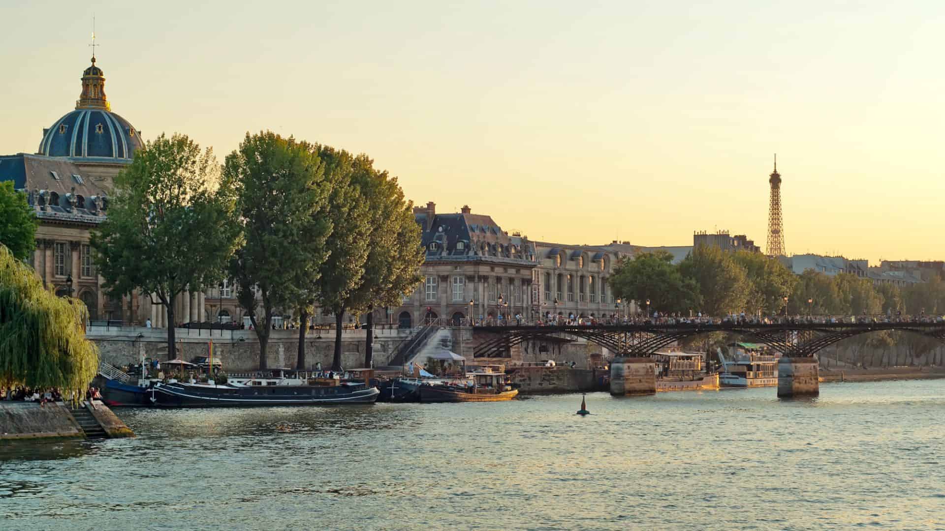 Seine River at sunset with boats, trees, Paris buildings, Pont des Arts, and Eiffel Tower in the background.