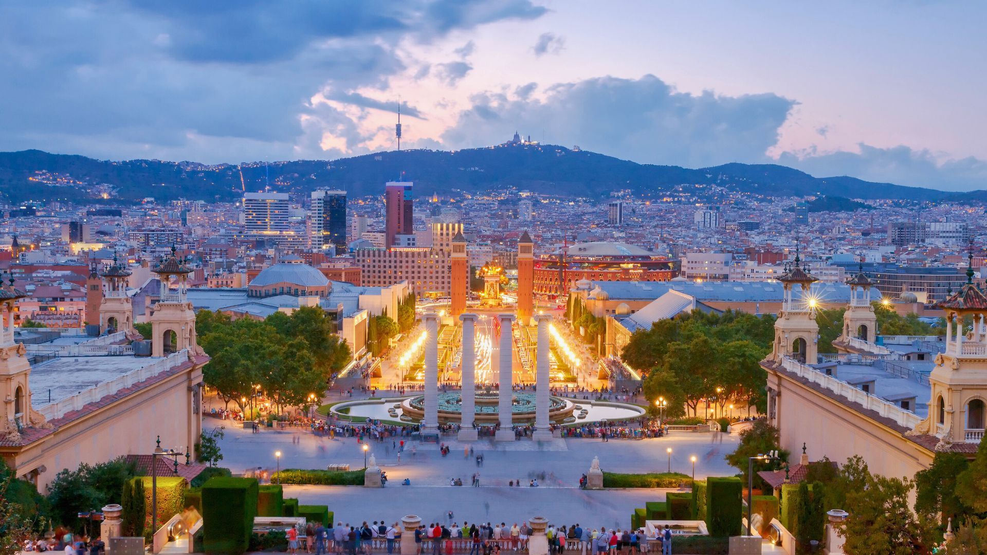 View of a cityscape at sunset with illuminated fountains, columns, and distant mountains in the background.